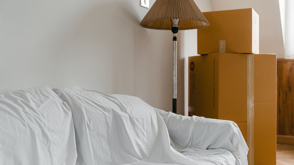 Packed cardboard boxes and a sheet-covered sofa with a floor lamp in a light, spacious room